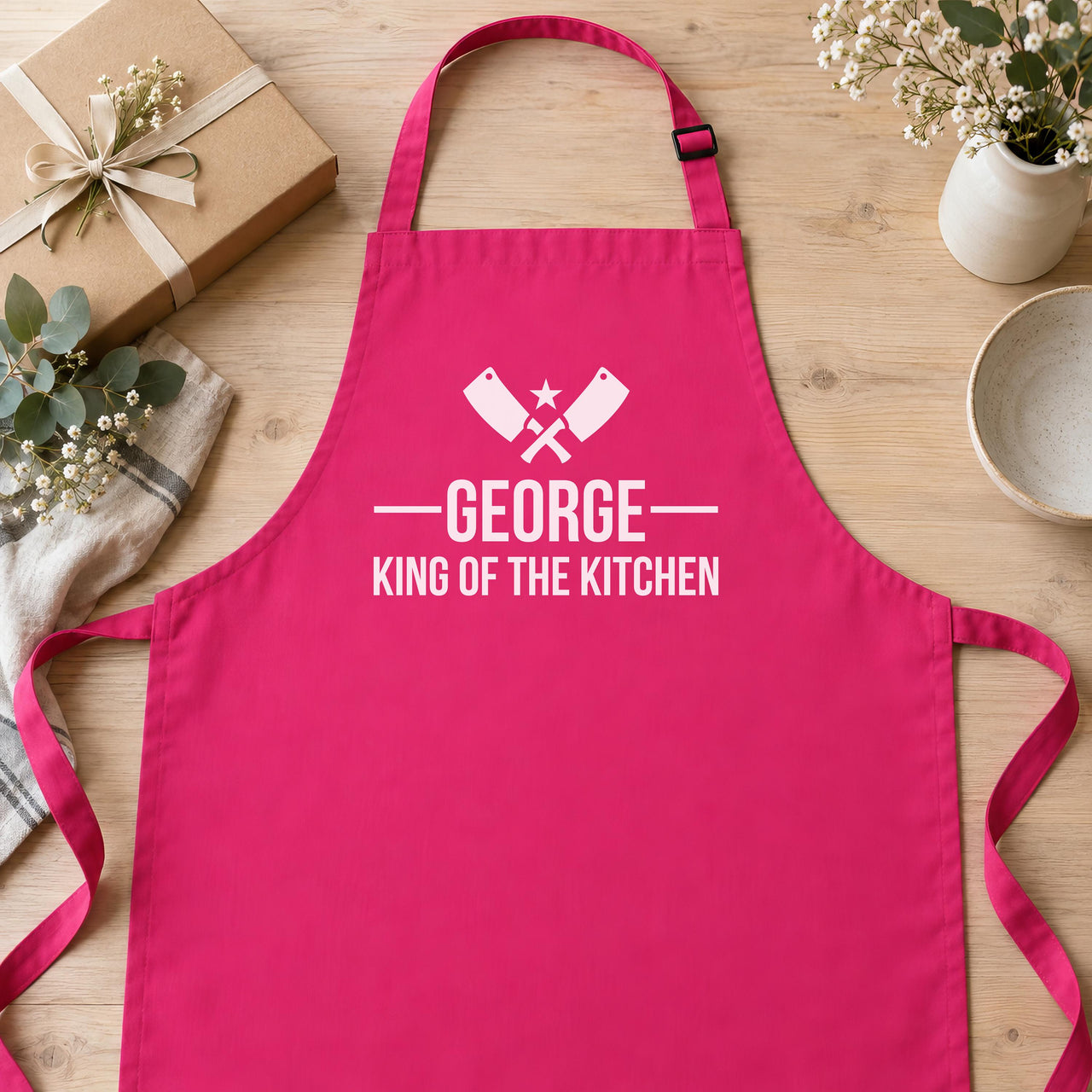Pink apron flat lay with gift box and flowers, 'GEORGE King of the Kitchen' and crossed cleavers - personalised apron