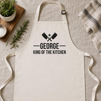 Natural cotton apron laid flat on linen, black 'GEORGE King of the Kitchen' print with cleavers - personalised apron