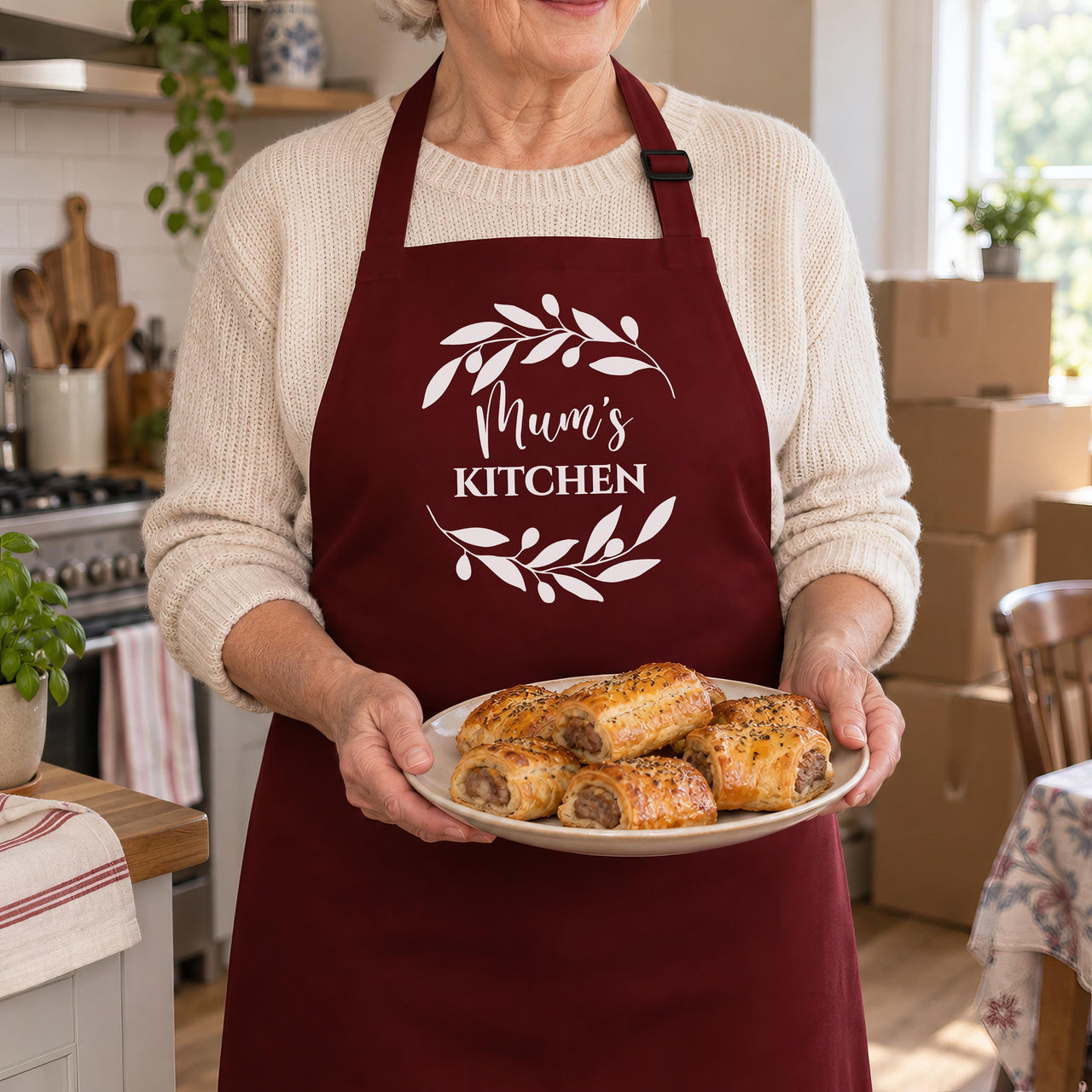 Burgundy apron with Mum's Kitchen inside a leaf wreath, worn by a woman holding sausage rolls