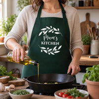 Green Mum's Kitchen apron with a leaf wreath, worn while pouring olive oil into a pan