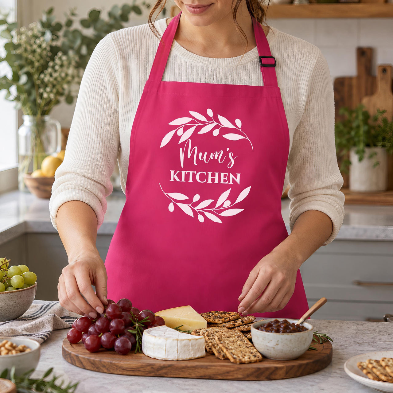Pink Mum's Kitchen apron with a leaf wreath, worn while laying out a cheese board
