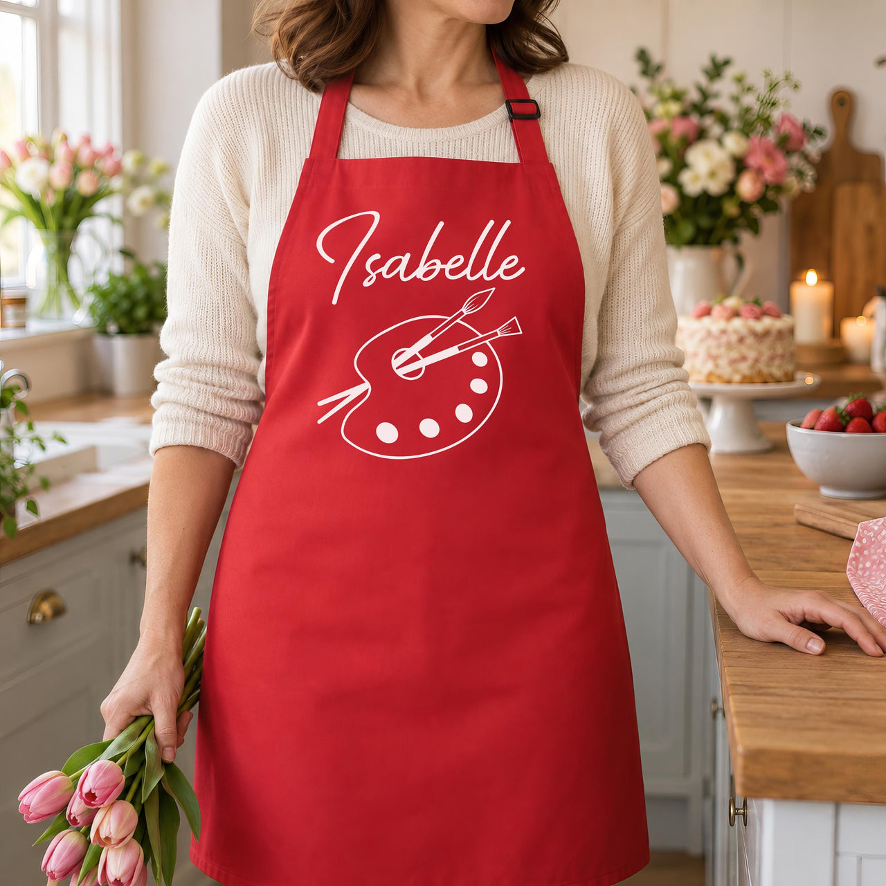 Red apron with white 'Isabelle' and paint palette design, holding pink tulips - personalised apron