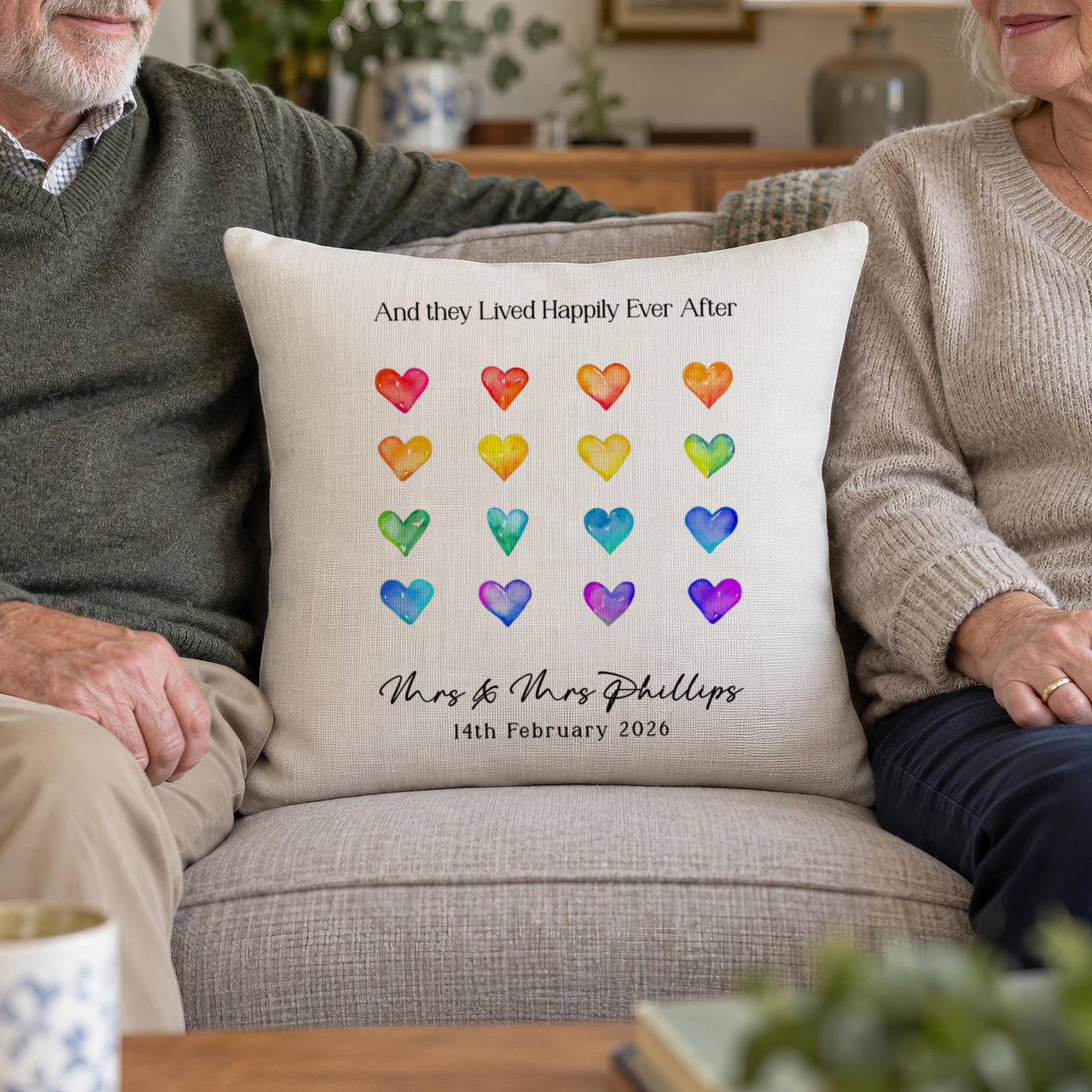 Rainbow hearts wedding cushion 'Mrs & Mrs Phillips' on a sofa between an older couple - personalised cushion