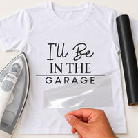 Mockup: white tee reading 'I'll Be IN THE GARAGE' in mixed script and capitals, iron and black vinyl roll nearby