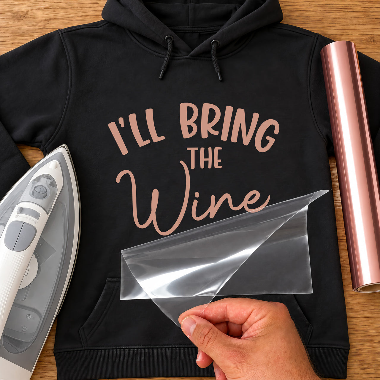 Black hoodie with rose gold 'I'LL BRING THE Wine' lettering, carrier film being peeled off - iron-on vinyl transfer