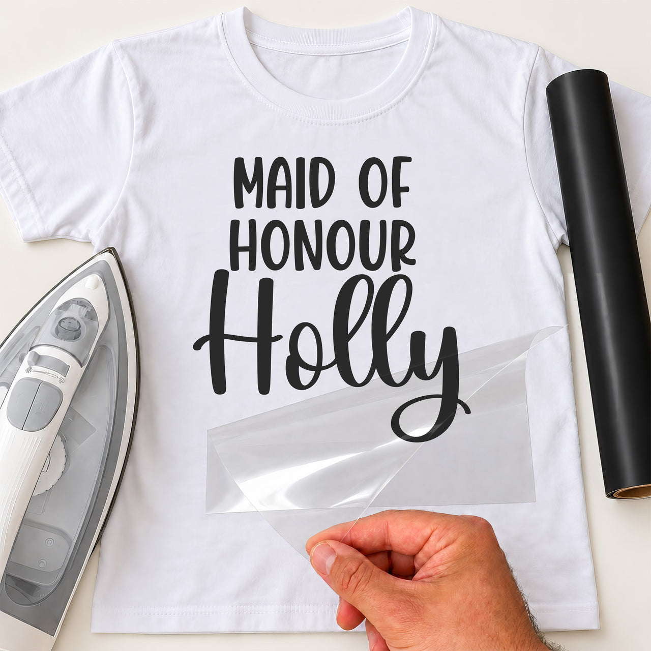 Maid of Honour iron-on transfer: black MAID OF HONOUR Holly on a white T-shirt, film being peeled