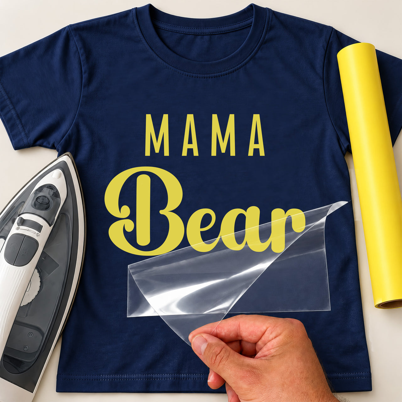 Navy T-shirt mockup showing yellow "MAMA" over a script "Bear", with an iron and yellow vinyl roll