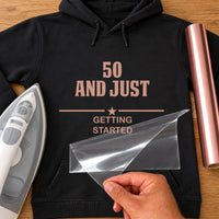 Milestone Birthday glitter iron-on: rose gold 50 AND JUST GETTING STARTED on a black hoodie, film being peeled
