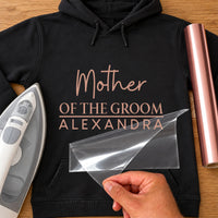 Black hoodie mockup with rose gold 'Mother OF THE GROOM ALEXANDRA', iron and rose gold vinyl roll beside it