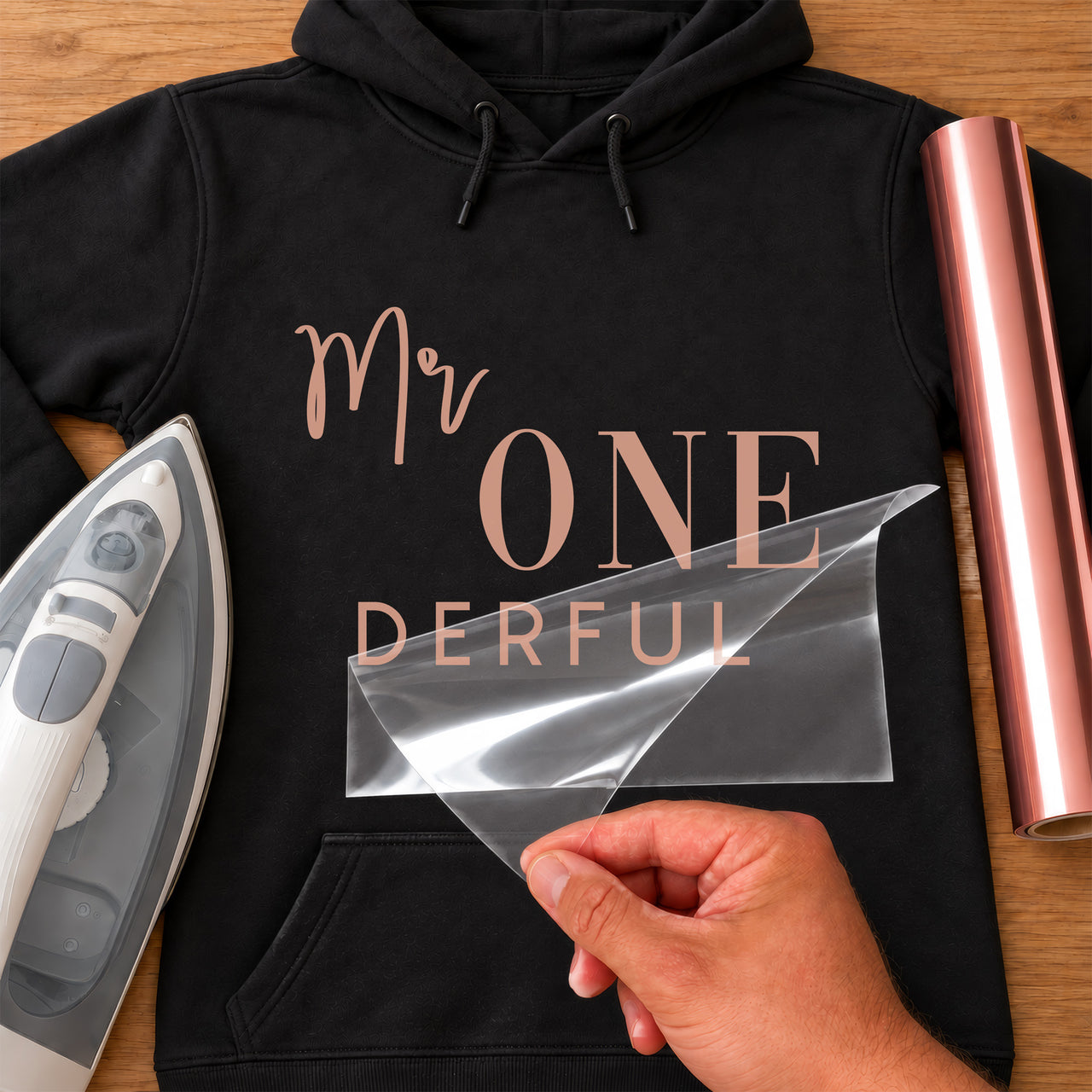 Mockup: black hoodie with rose gold vinyl 'Mr ONE DERFUL' first birthday wording, iron and vinyl roll beside
