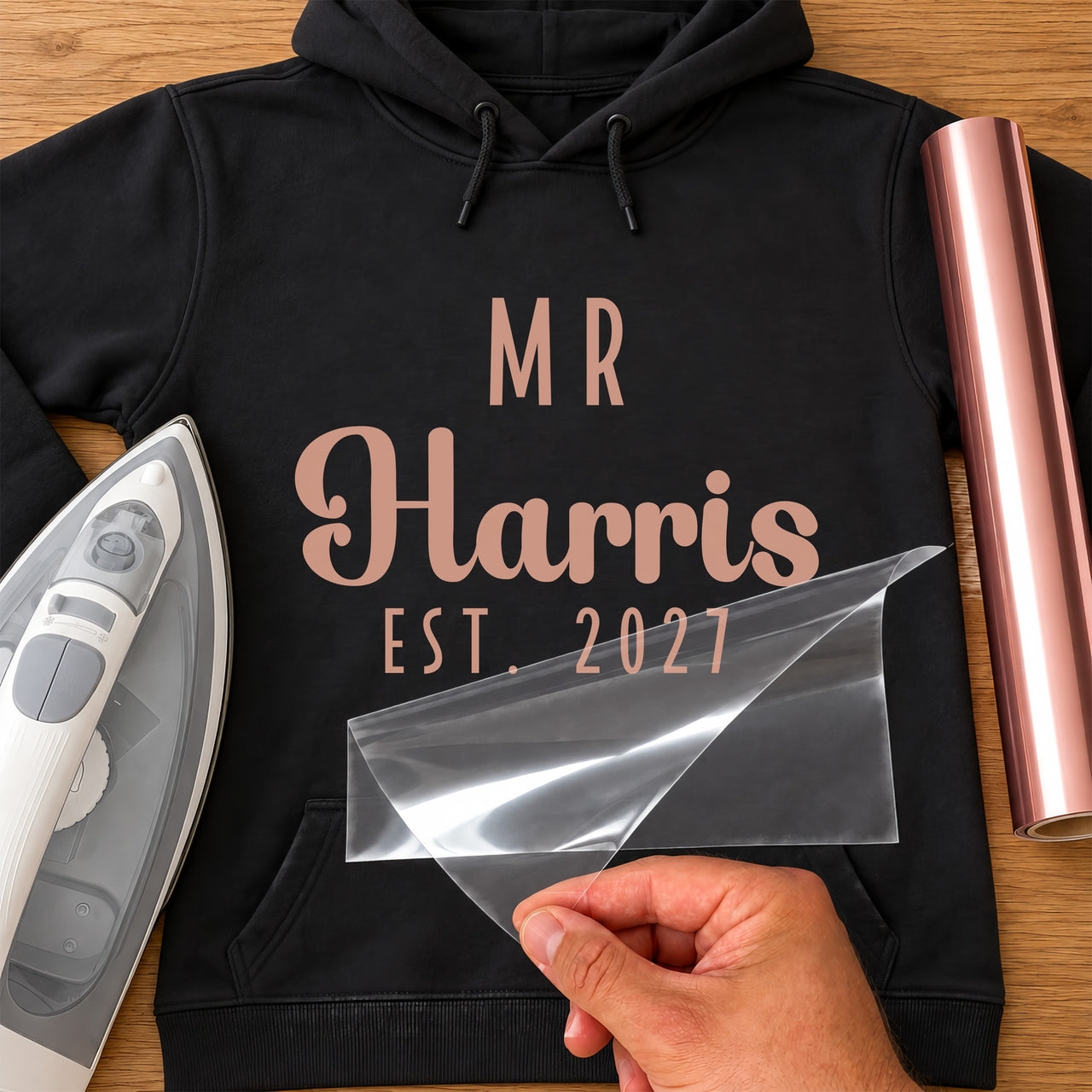 Mr Surname Est. iron-on transfer: rose gold MR Harris EST. 2027 on a black hoodie, film being peeled
