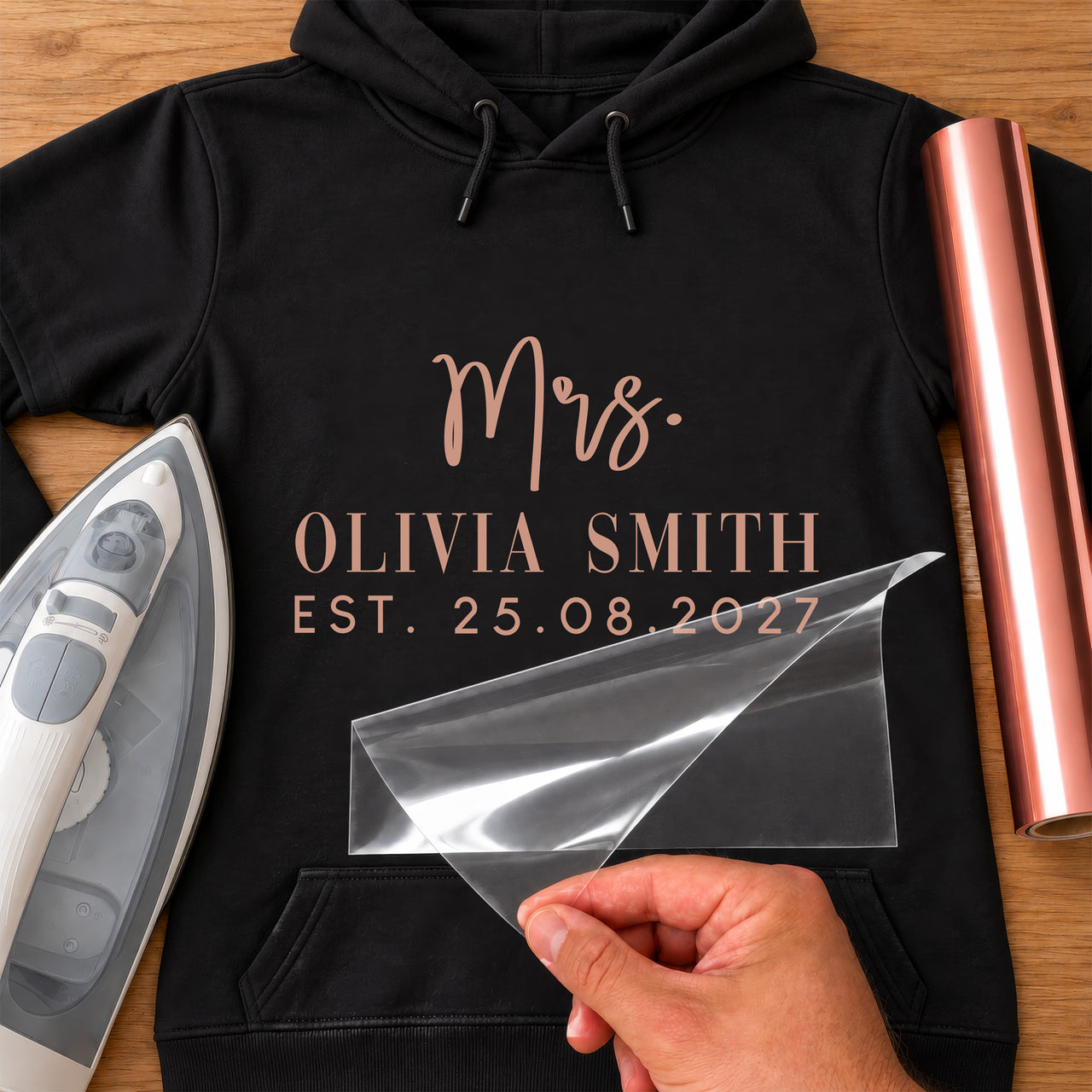 Black hoodie with rose gold 'Mrs.' 'OLIVIA SMITH' 'EST. 25.08.2027', carrier film being peeled - iron-on vinyl transfer