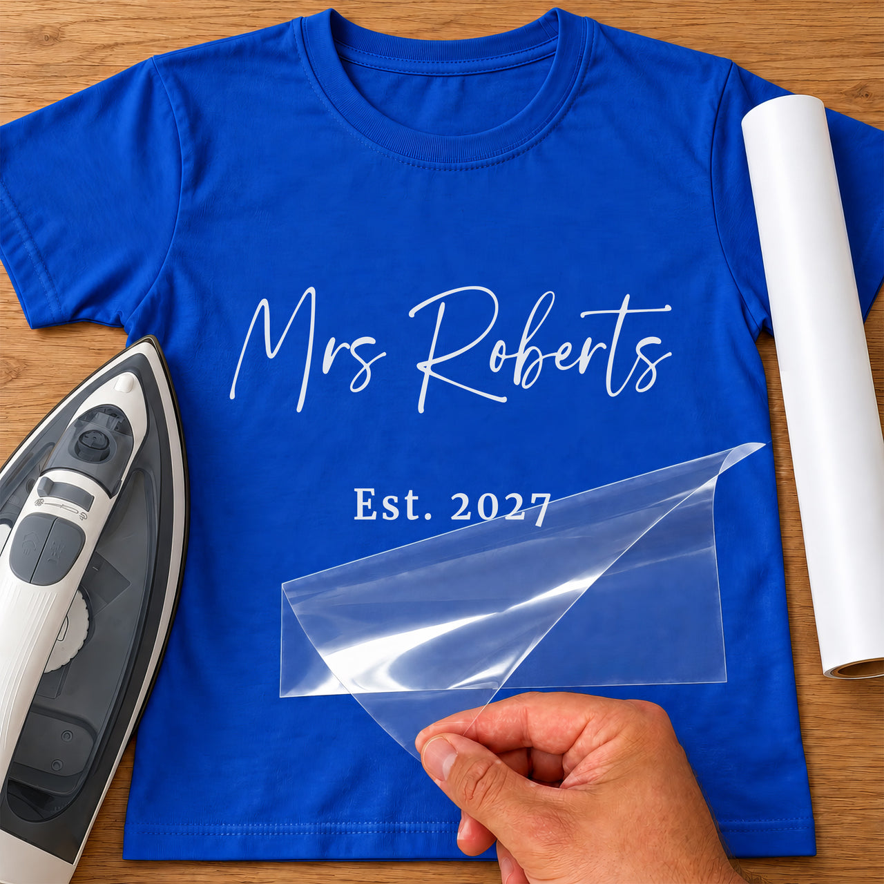 Blue T-shirt with white 'Mrs Roberts' and 'Est. 2027' lettering, carrier film being peeled off - iron-on vinyl transfer