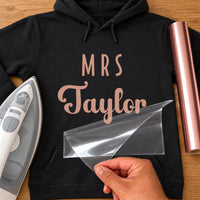 Black hoodie with rose gold 'MRS Taylor' iron-on vinyl transfer, carrier sheet being peeled off, iron and vinyl roll beside