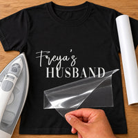 Mockup of black T-shirt with white script 'Freya's' over serif 'HUSBAND', clear film peeled back by hand