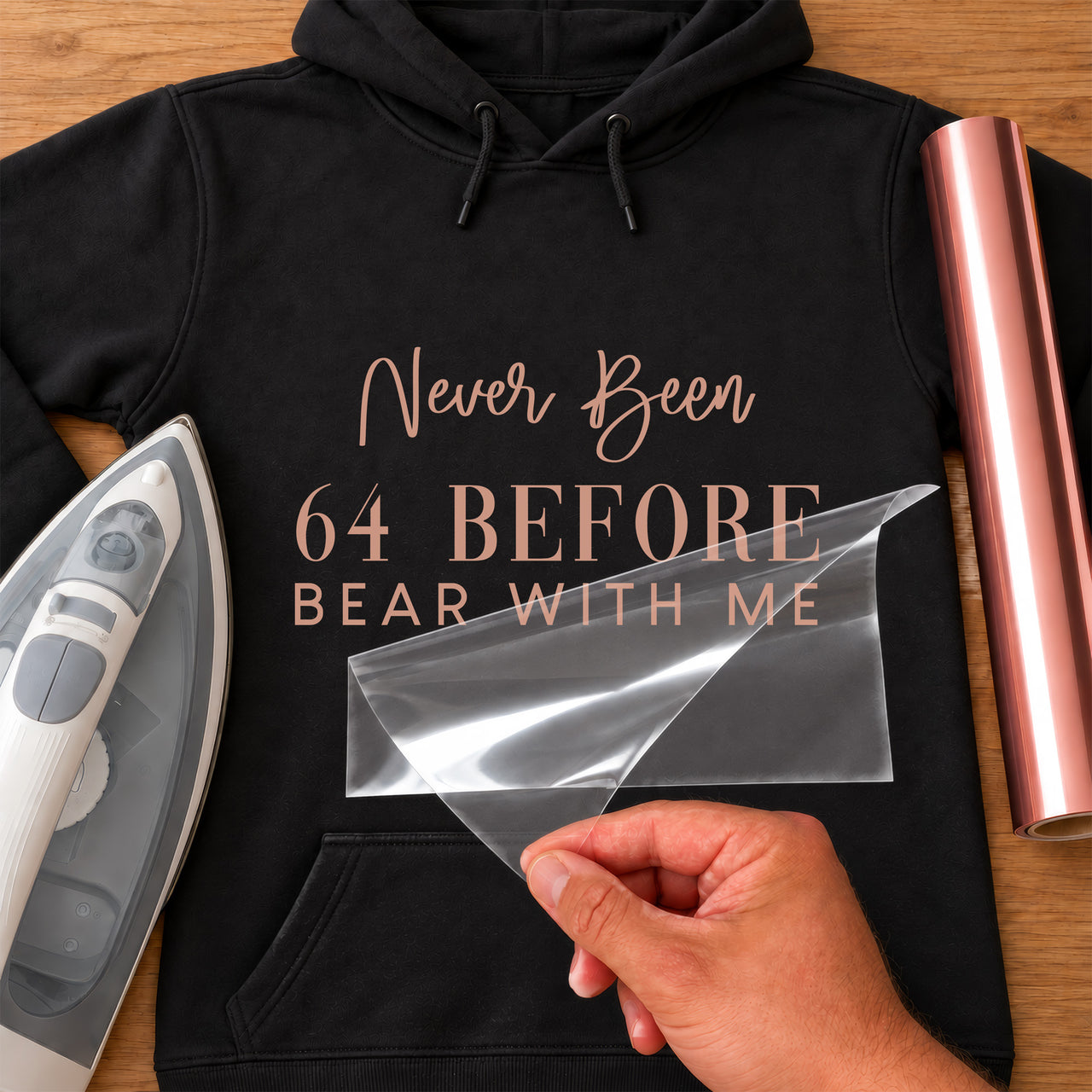 Never Been 64 birthday iron-on transfer: rose gold Never Been 64 BEFORE BEAR WITH ME on a black hoodie, film being peeled