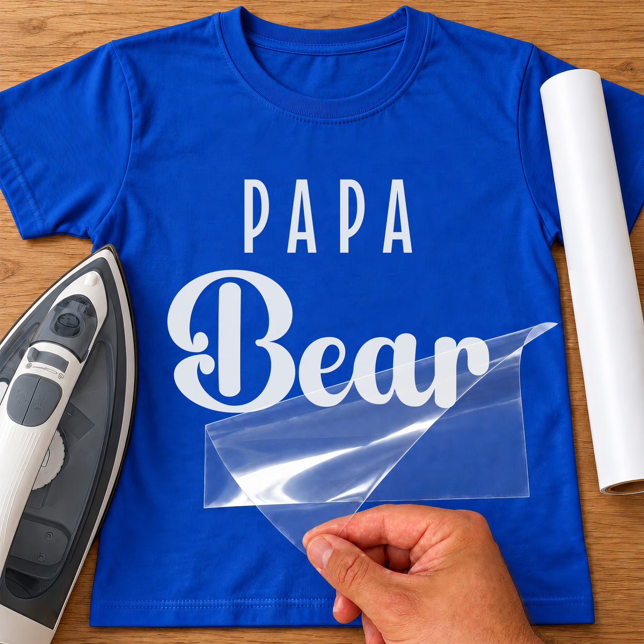 Mockup: blue T-shirt with white 'PAPA' and script 'Bear', clear carrier sheet being peeled, iron and white vinyl roll