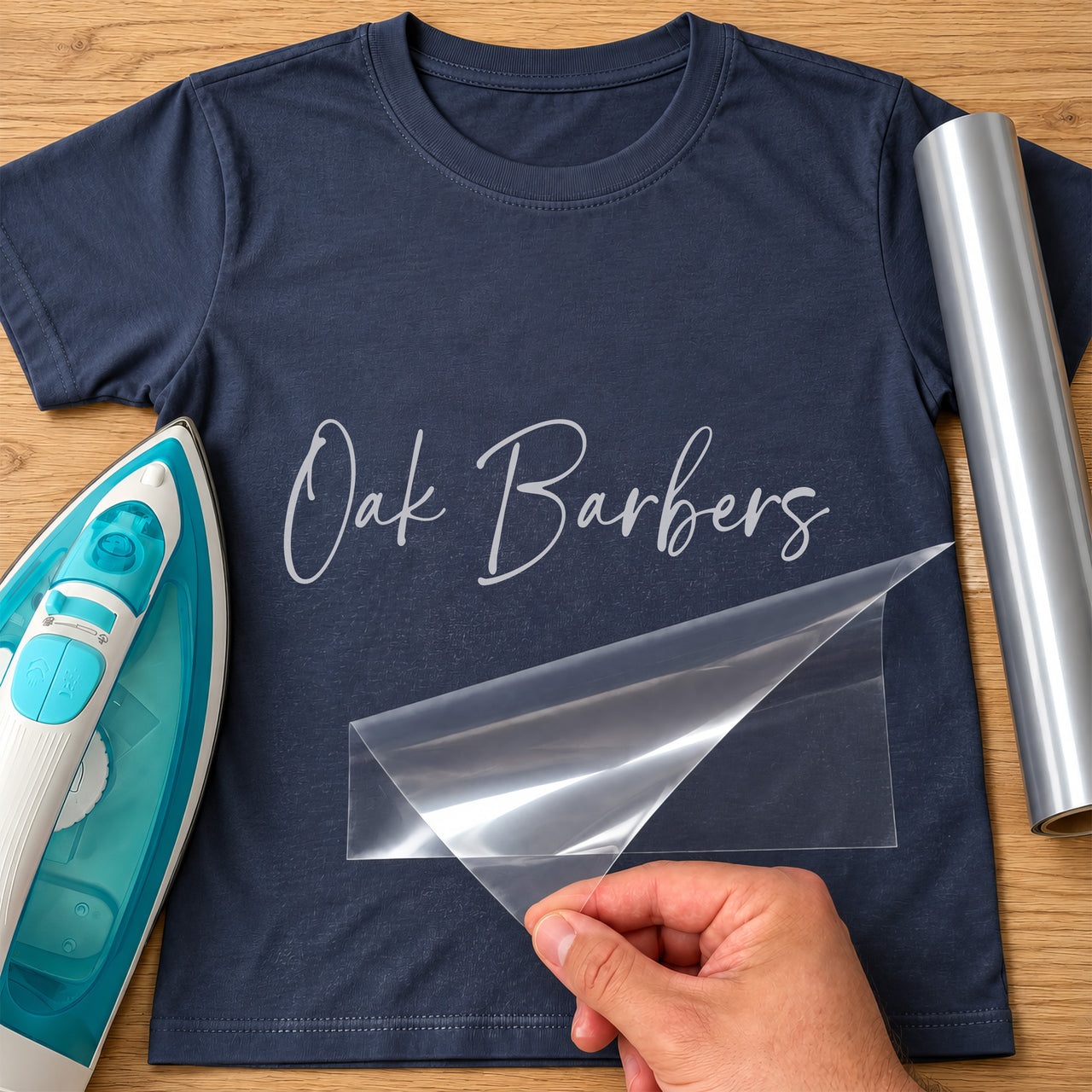 Personalised Barber Name iron-on transfer: silver script Oak Barbers on a navy T-shirt, film being peeled