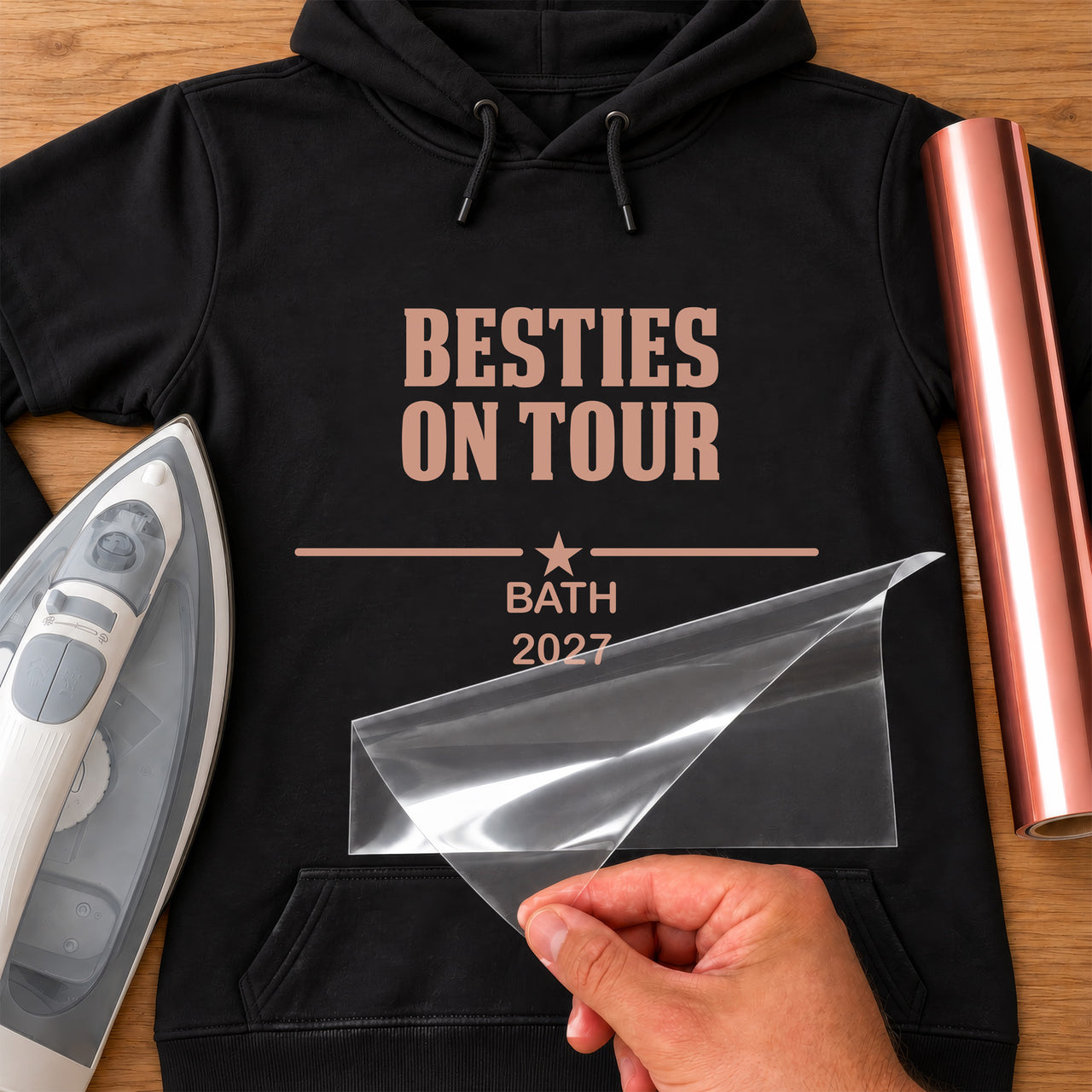 Besties Trip iron-on transfer: rose gold BESTIES ON TOUR BATH 2027 on a black hoodie, film being peeled