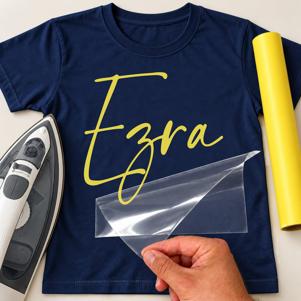 Personalised Childminder Name iron-on transfer: yellow script Ezra on a navy T-shirt, film being peeled