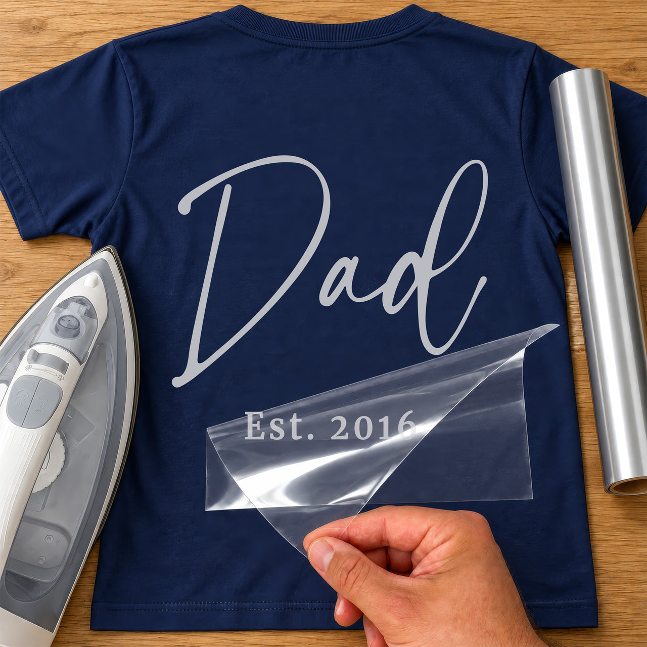 Dad Est. Year iron-on transfer: silver script Dad with Est. 2016 below on a navy T-shirt, film being peeled