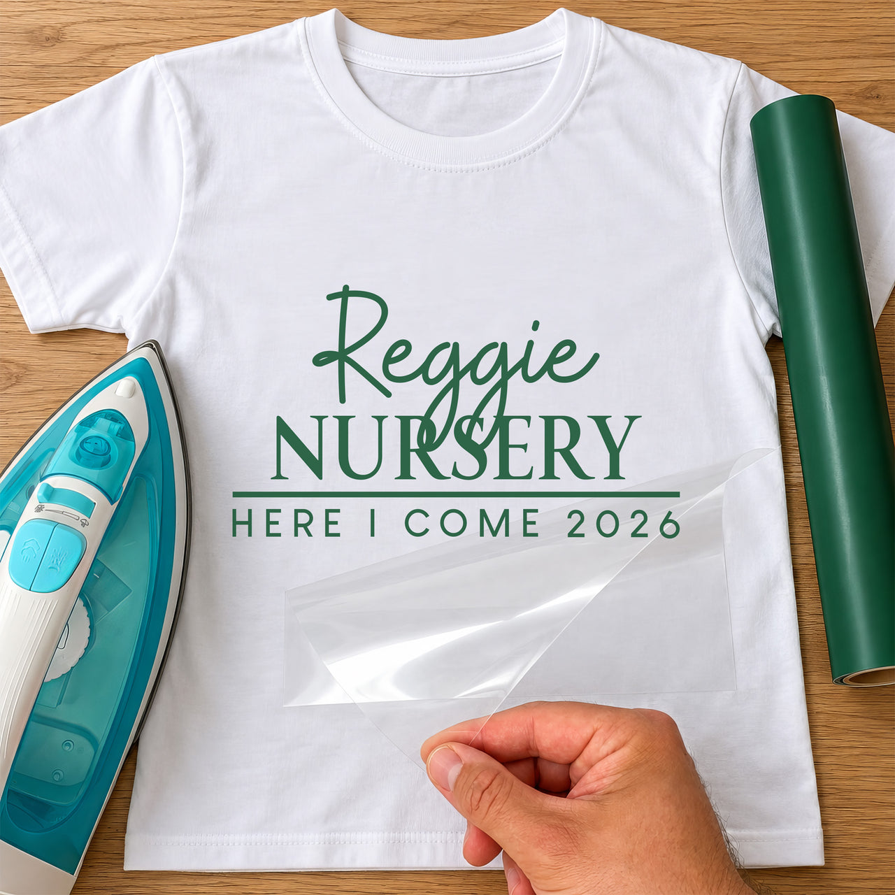 First Day of Nursery iron-on transfer: green Reggie NURSERY HERE I COME 2026 on a white T-shirt, film being peeled