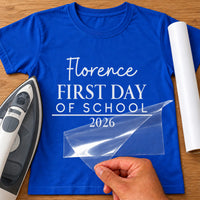 First Day of School iron-on transfer: white Florence FIRST DAY OF SCHOOL 2026 on a royal blue T-shirt, film being peeled