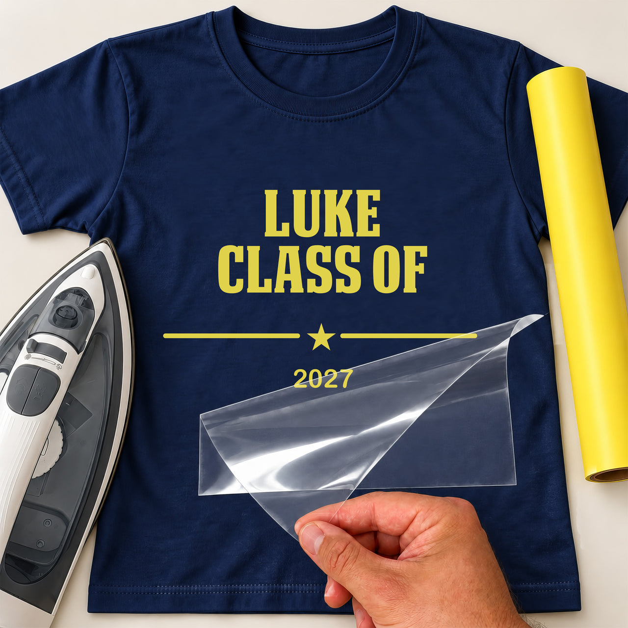 Graduation 2026 iron-on transfer: yellow LUKE CLASS OF 2027 on a navy T-shirt, film being peeled