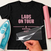 Lads Holiday iron-on transfer: pink LADS ON TOUR KIERAN 2027 on a black T-shirt, film being peeled