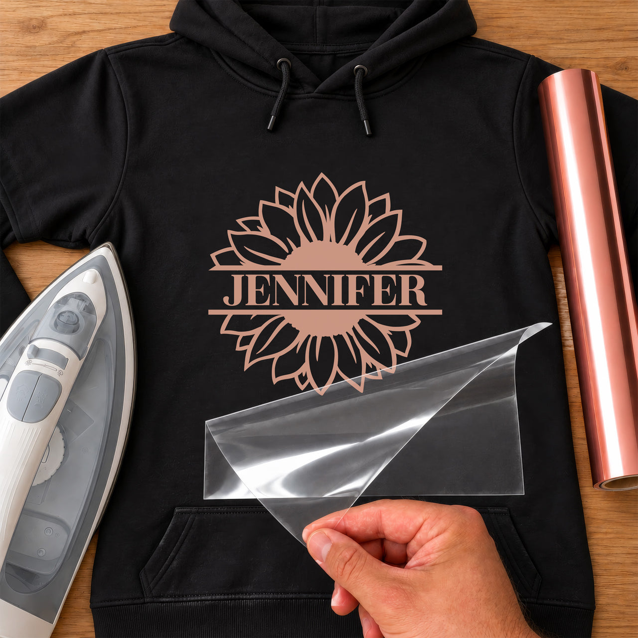 Black hoodie with rose gold sunflower and 'JENNIFER' split name, carrier film being peeled off - iron-on vinyl transfer