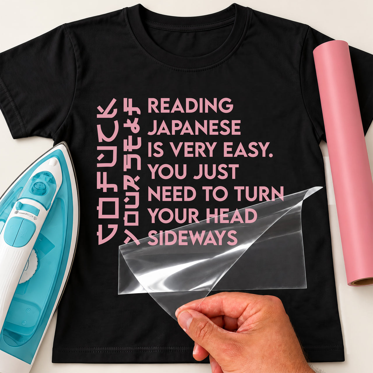 Black T-shirt with pink sideways joke lettering transfer, carrier sheet being peeled near an iron