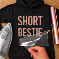 Black hoodie with rose gold 'SHORT BESTIE' iron-on vinyl transfer, carrier sheet being peeled off, iron and vinyl roll