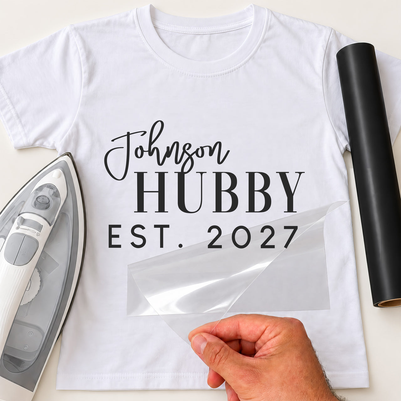 White T-shirt mockup with script 'Johnson', serif 'HUBBY' and 'EST. 2027', clear transfer film lifted by hand