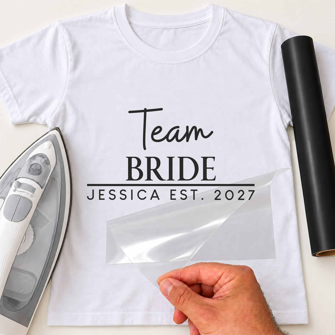 White tee reading 'Team BRIDE' and 'JESSICA EST. 2027' in black, hen do iron-on transfer with film half peeled