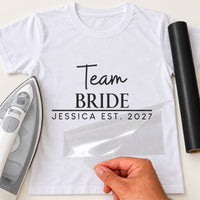 White tee reading 'Team BRIDE' and 'JESSICA EST. 2027' in black, hen do iron-on transfer with film half peeled