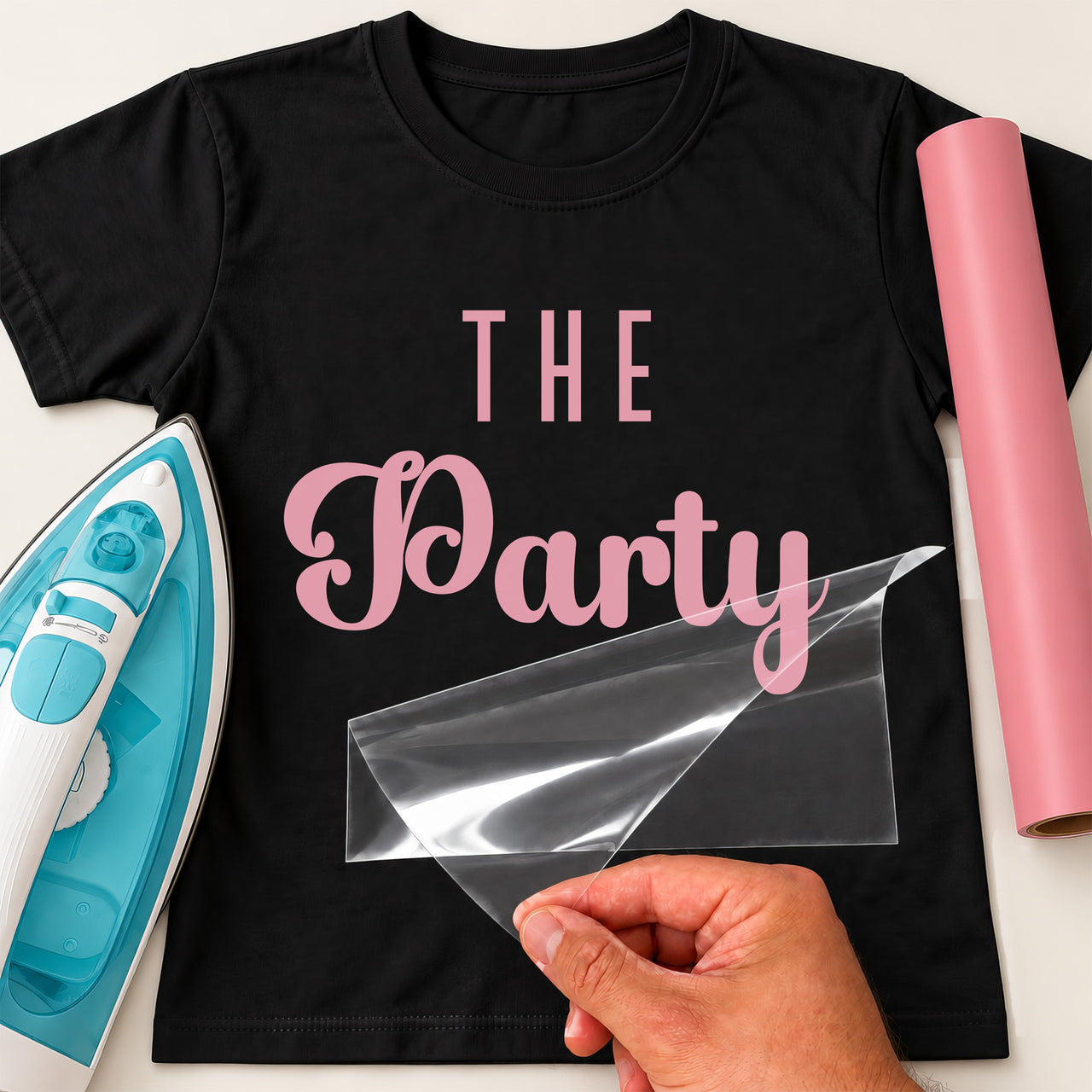 Black T-shirt with pink 'THE Party' iron-on vinyl transfer, carrier sheet being peeled off, iron and pink vinyl roll