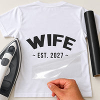 Mockup: white tee with arched bold 'WIFE' and '~ EST. 2027 ~' below, clear carrier sheet peeled, iron and black vinyl
