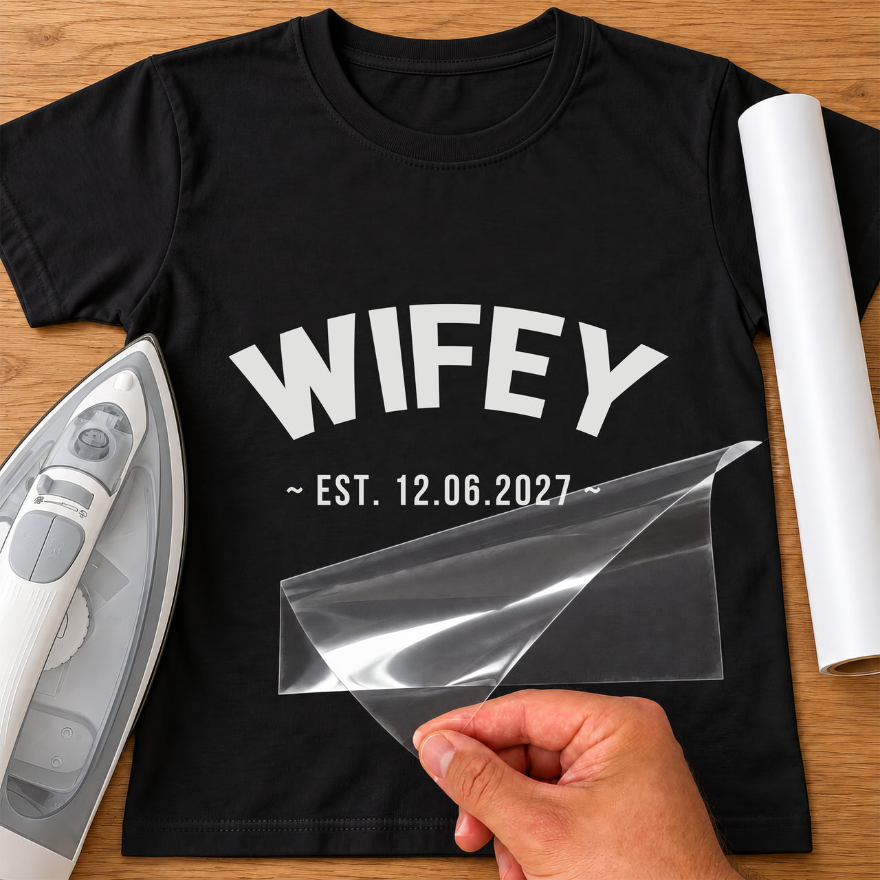 Wifey date iron-on transfer: white WIFEY EST. 12.06.2027 on a black T-shirt, film being peeled