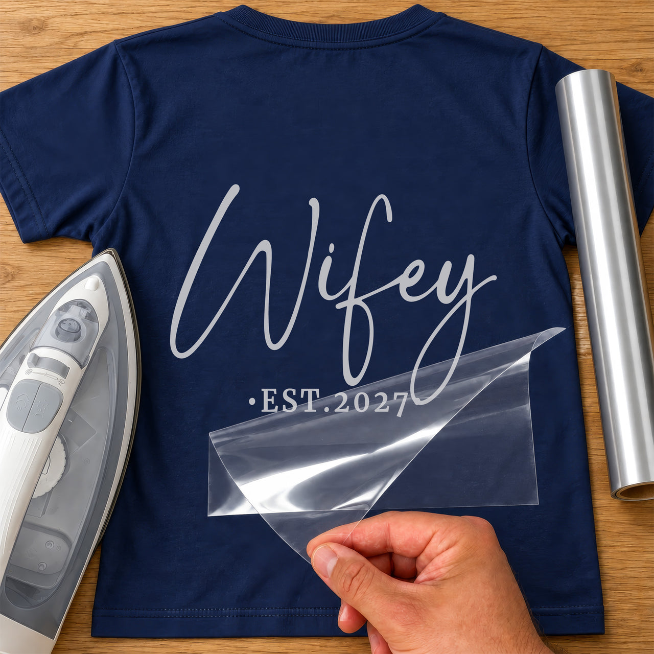 Back of a navy T-shirt with silver script 'Wifey' and '•EST.2027', carrier film being peeled off - iron-on vinyl transfer
