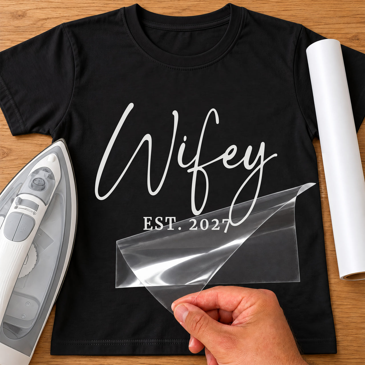 Black T-shirt with white script 'Wifey' and 'EST. 2027', carrier film being peeled off - iron-on vinyl transfer