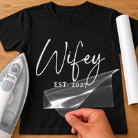 Black T-shirt with white script 'Wifey' and 'EST. 2027', carrier film being peeled off - iron-on vinyl transfer