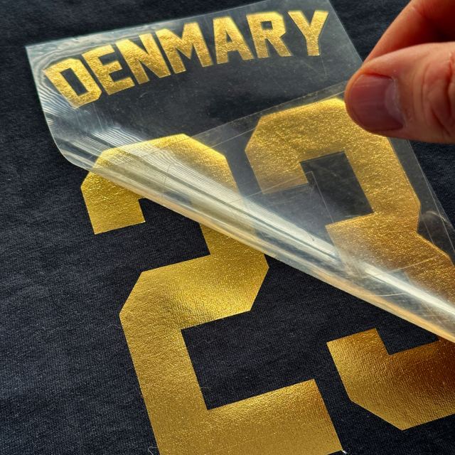 Clear sheet peeled off a gold metallic DENMARY 23 on a navy T-shirt