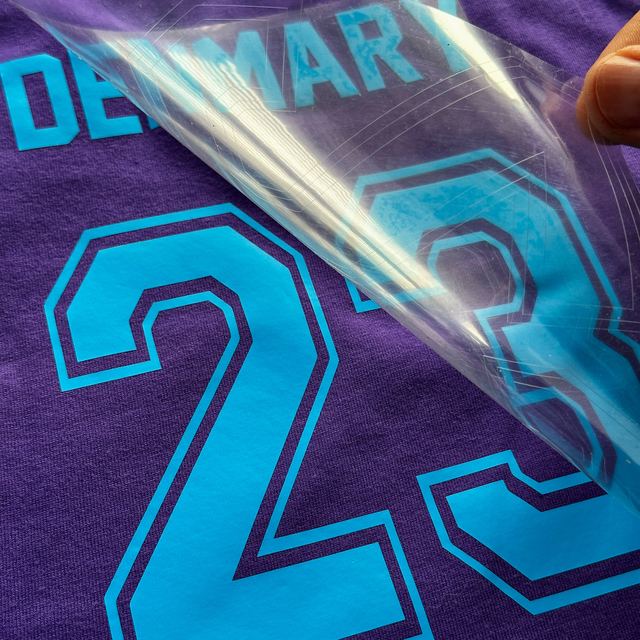 Clear sheet peeled off a neon blue DENMARY 23 on a purple T-shirt