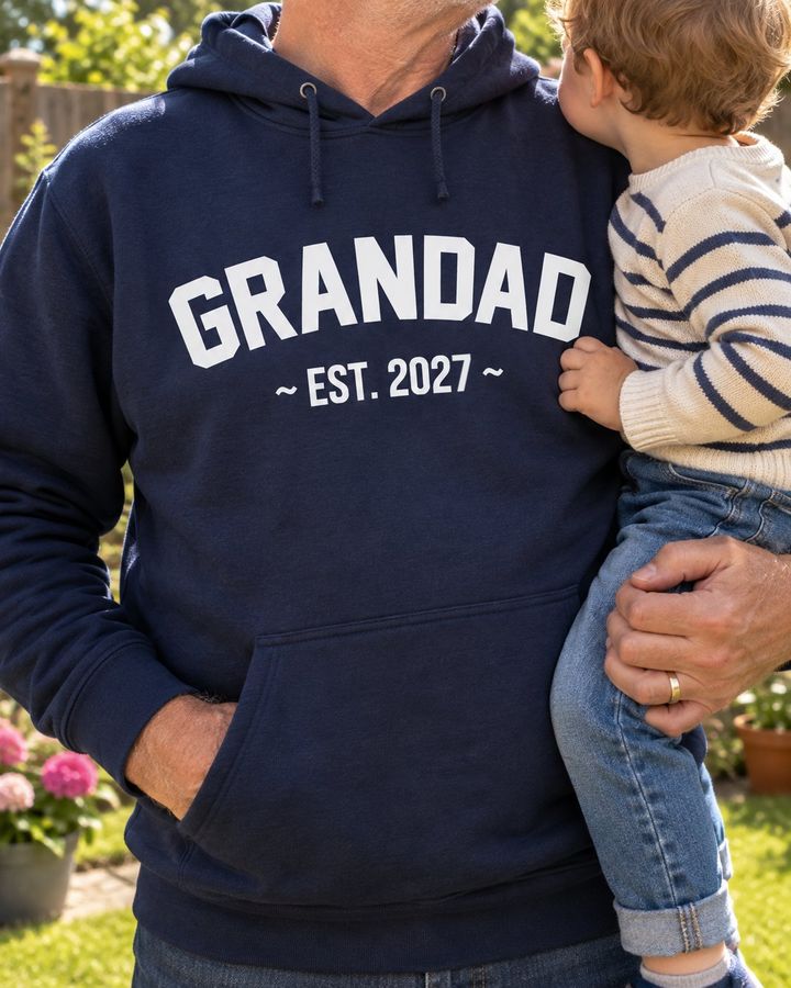 Grandad in a navy hoodie with GRANDAD EST. 2027 in white, holding a toddler in the garden